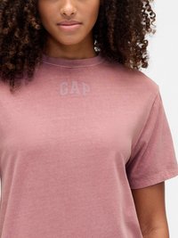 Woman with curly hair wearing a faded pink GAP T-shirt, shown from the shoulders to mid-torso against a plain background.