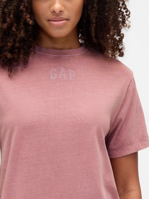 Woman with curly hair wearing a faded pink GAP T-shirt, shown from the shoulders to mid-torso against a plain background.