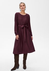 Mauve sukienka z długim rękawem, z wcięciem w talii i detalem w postaci kokardy; miękka, gładka tkanina; długość do kostek; modelka zestawiła ją z brązowymi, sięgającymi kolan botkami.