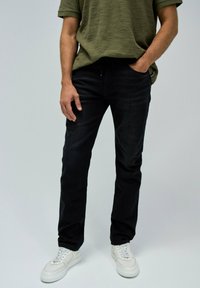 Jeans de mezclilla negra con un corte ajustado, que presentan una cintura regular. Combinados con una camisa de color verde oliva texturizada y zapatillas blancas.