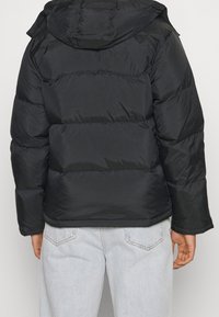 Doudoune noire avec capuche, dotée de sections matelassées horizontales, de poignets élastiques et d'un ourlet cintré pour la chaleur et le confort.