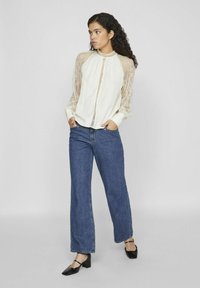 Blusa de marfil con largas mangas de encaje y cuello alto, con botones en la parte delantera; combinada con unos vaqueros de pierna ancha de mezclilla azul y zapatos de tacón negros.