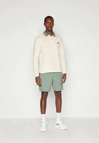 Bež dug rukav polo majica s ovratnikom i sivim detaljima, u kombinaciji sa svijetlozelenim kratkim hlačama. Bijele tenisice i čarape do gležnjeva upotpunjuju izgled.