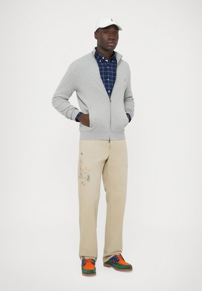 Világosszürke cipzáras kardigán, tengerészkék kockás ing, bézs nadrág hímzett mintával, és zöld, narancs, valamint kék színekben pompázó színes cipők.