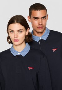 Navy long-sleeve polo shirts with light denim collars, featuring a red triangular logo badge on the left side. Smooth texture, casual fit.