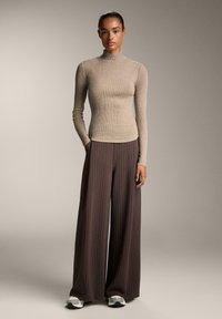 Kvinde står iført beige ribbestrikket turtleneck, brune bukser med brede striber og hvide sneakers foran en neutral baggrund.
