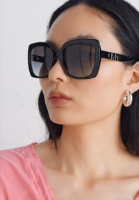 Suur must ruudukujuline prillid gradient läätsedega ja sleek raamiga. Tekstureeritud must kõrvarõngas täiendab välimust, valge tausta ees.