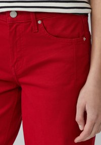 Un pantalon en coton rouge présente une coupe droite, des poches avant et des accents en métal argenté avec des détails de couture visibles. Associé à un haut rayé.