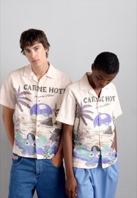 Short-sleeve, cream button-up shirt featuring tropical print with palm trees, a purple sun, and blue accents. Worn by two models.