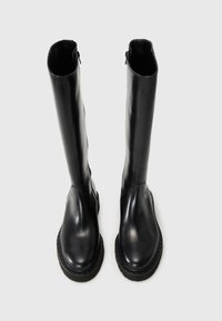 Botas de cuero negras hasta la rodilla con una textura suave, puntera redondeada y suela de goma gruesa. Cierre con cremallera lateral.