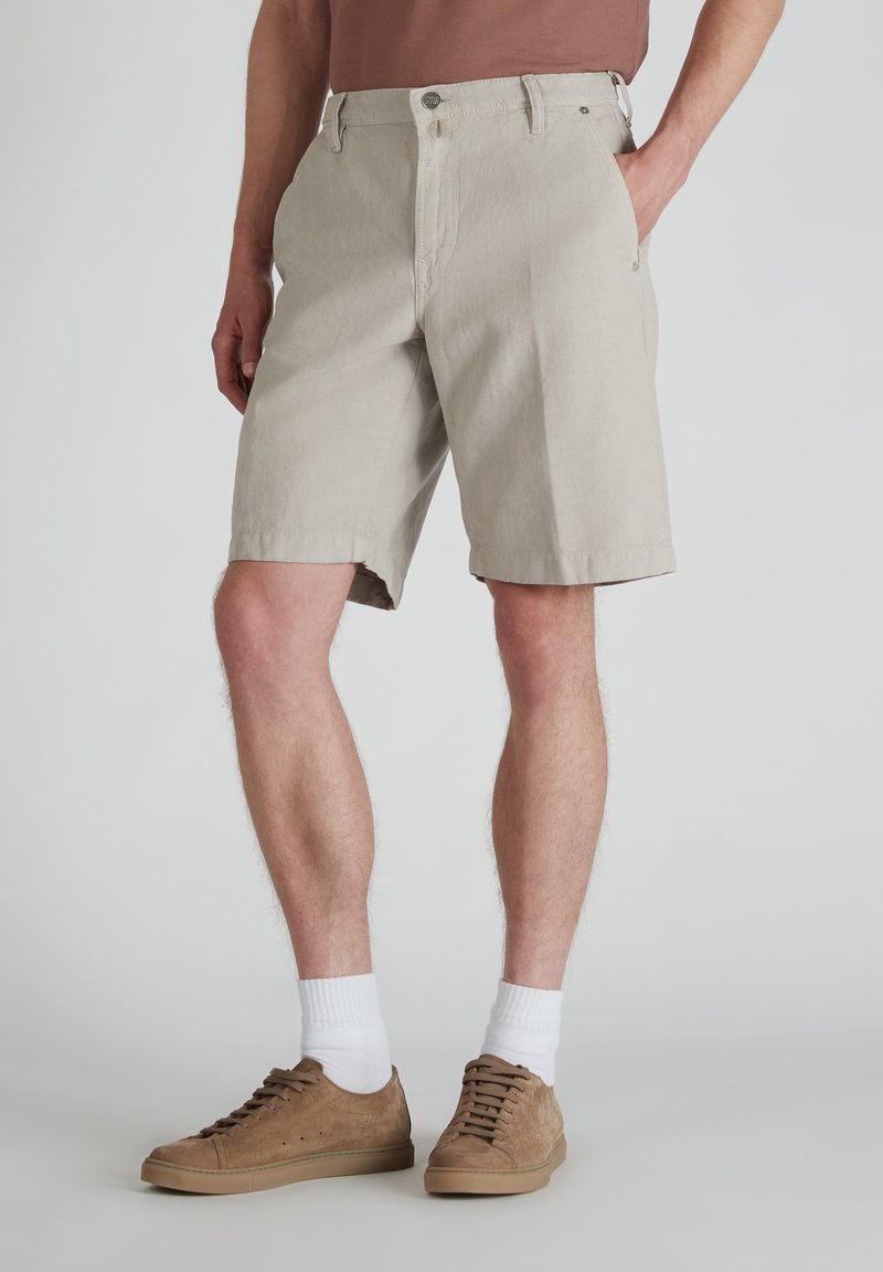 Pantalones cortos de algodón beige con un corte recto, dos bolsillos frontales y un cierre de botón. Combinados con zapatos de ante marrones y calcetines blancos.