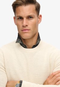 Cremfarbener Pullover mit Textur über einem grün-schwarz karierten Hemd, mit runden Ausschnitt und subtilen Rippendetails.