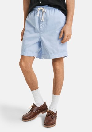 Shorts légers bleu clair avec une taille élastique et un cordon de serrage. Portés avec des chaussures bateau en cuir marron et des chaussettes blanches.