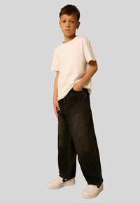 Boy standing with hand in pocket wearing white t-shirt, loose black jeans, and white sneakers against plain gray background.