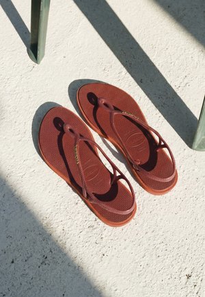 Par brune Havaianas-sandaler med tæseremme placeret på tekstureret hvidt gulv, skygger kastet diagonalt i nærheden.