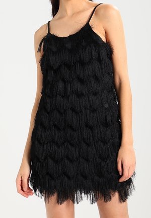 Femme portant une mini-robe noire sans manches avec des détails de franges superposées et des fines bretelles.