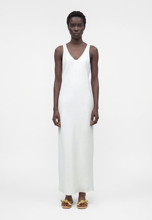 Wysoka osoba z krótkimi kręconymi włosami, ubrana w białą, długą sukienkę bez rękawów oraz żółte sandały z kokardkami, stojąca na jednolitym tle.