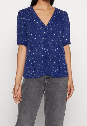Mujer con blusa azul de manga corta abotonada con un pequeño estampado floral y vaqueros grises de talle alto.