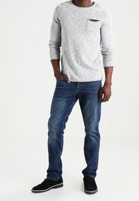 Camicia a maniche lunghe grigia chiaro con tasca sul petto, abbinata a jeans blu scuro e sneaker nere. Presenta una vestibilità casual e un tessuto testurizzato.