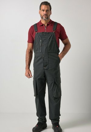 Mann mit kurzen dunklen Haaren und Bart, trägt schwarze Arbeitsoveralls und ein rotes kurzärmeliges Poloshirt, steht vor einem schlichten hellen Hintergrund.