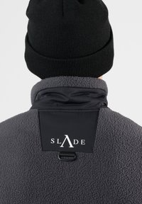 Giacca in pile grigia con colletto in nylon nero, dotata di un patch con logo e di un dettaglio a ciclo sul retro. Cappellino in maglia nero indossato sopra.