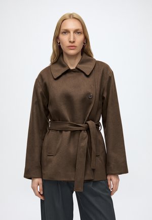 Femme aux longs cheveux blonds porte un manteau marron ceinturé avec un large col et un bouton visible, se tenant devant un fond clair uni.