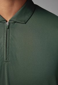 Tamnozelena polo majica s teksturiranim uzorkom, koja ima zatvaranje na pola munje i ravni ovratnik. Materijal izgleda glatko i lagano.