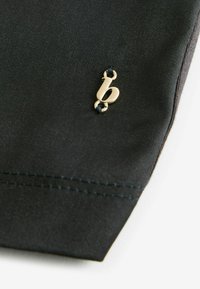 Černý látkový sáček s malým zlatým emblémem písmene "b" a kontrastním modrým prošíváním podél švu. Hladká textura, elegantní design.