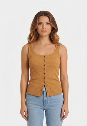 Donna con capelli castano mossi che indossa un top cammello senza maniche con bottoni e jeans azzurri, in piedi contro uno sfondo bianco.