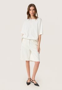 Hvid oversized T-shirt og knælange shorts, begge i et blødt stof. Sorte spidse sko med ankelremme. Enkel, ren design.