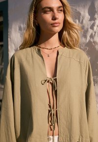 Svetloolivovo zelená voľne strihaná bunda so zaväzovaním vpredu a dlhými rukávmi. Vyrobená z mäkkej tkaniny s uvoľnenou siluetou.