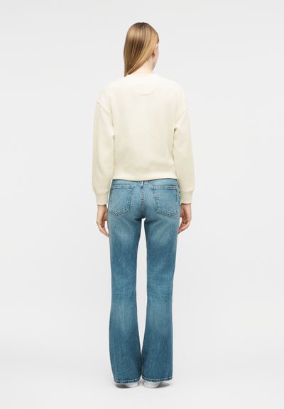 Γυναίκα με ίσια ξανθά μαλλιά φοράει κρεμ φούτερ και μπλε τζιν καμπάνα, στέκεται γυρισμένη ανάποδα σε απλό φόντο.