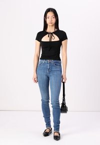 Fekete, testhez simuló top kivágott résszel és megkötős nyakkal, kék skinny farmerral párosítva. A modell fekete lapos cipőt visel, és egy kis fekete táskát tart a kezében.