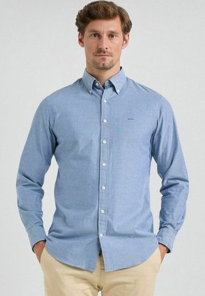 Man draagt lichtblauw overhemd met knopen en beige broek, staat met handen in zakken tegen een effen achtergrond.