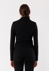 Blazer nero aderente con maniche lunghe, texture liscia, spalline discrete e colletto a scollatura. Design sartoriale con schiena liscia.