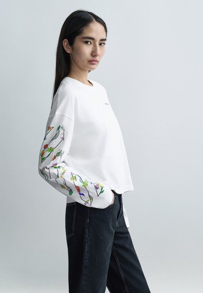 White long-sleeve t-shirt with floral embroidery in colors on sleeves, paired with dark jeans; features a round neckline and relaxed fit.