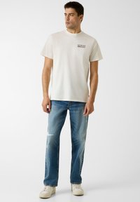 T-shirt couleur crème avec le logo "GUESS" sur la poitrine, associée à un jean bleu délavé aux détails usés, porté avec des baskets blanches.