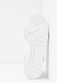 Weißer Sneaker mit kompliziertem, strukturiertem Muster an der Sohle; goldene Akzente an den Seiten; mit "GUESS"-Logo nahe der Ferse; glattes, gummiartiges Material.