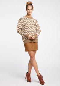 Pulover tricotat cu model zigzag maro, cu manșete împletite, purtat împreună cu o fustă din suede bej. Modelul poartă cizme scurte maro cu un design texturat.