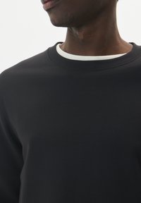 Čierna mikina s okrúhlym výstrihom, ktorá má kontrastný biely vnútorný golier. Hladká textúra, priliehavý strih a minimalistický štýl.