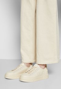 Sneakers beige con lacci e branding "LIU JO" indossati con pantaloni beige chiari a gamba larga su uno sfondo neutro.
