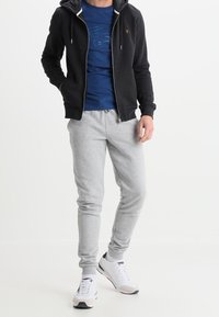 Felpa nera con zip, t-shirt grafica blu, pantaloni jogging grigi e scarpe sportive bianche. La felpa ha una tasca frontale e lacci.