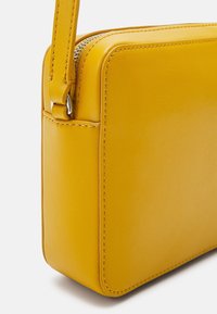 Gult læder crossbody-taske med rektangulær form, glat tekstur, lynlås-åbning og detaljerede stikninger langs kanterne.
