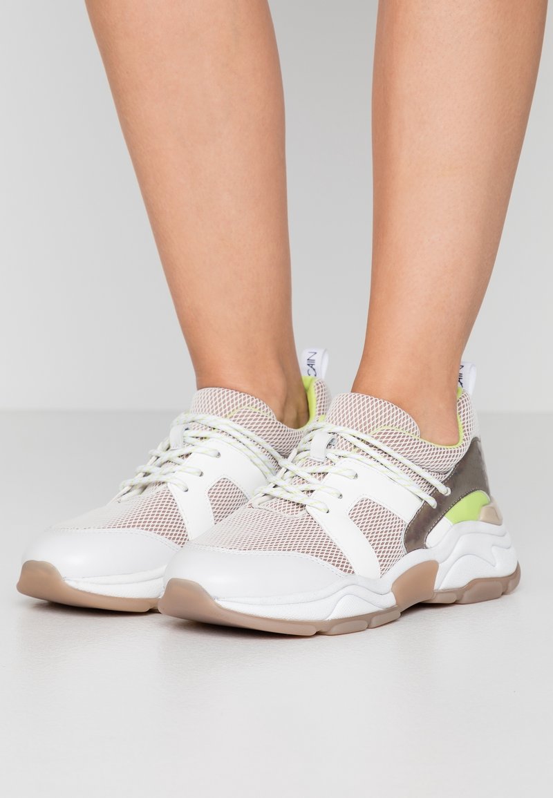 Baskets en maille blanche et gris clair avec semelles beige et accents vert clair portées pieds nus, vues de face et de côté.