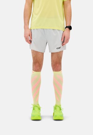 Mann trägt hellgraue Laufshorts, knielange Socken mit gelb-pinken Streifen, neongelbe Laufschuhe und ein gelbes Shirt.