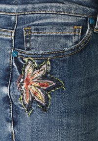 Džíny z denimu na modrém pozadí, s vícebarevným vyšívaným vzorem listu se flitry a kontrastním štepováním.