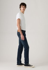 Tânăr îmbrăcat cu un tricou alb, blugi de culoare albastru închis și teniși negri, stând și uitându-se peste umăr pe un fundal simplu.