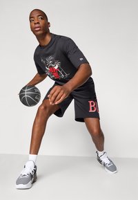 T-shirt negra com gráfico vermelho e design de palmeira, combinada com calções negros com um logo "BULLS" em vermelho. O modelo segura uma bola de basquete.