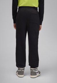 Calças de treino de algodão pretas com cintura elástica e um bolso na parte de trás, combinadas com ténis cinzentos de cano alto. A textura parece macia e confortável.