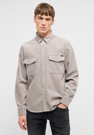 Ung mand står vendt fremad iført en grå kordfløjls-skjorte med knapper og to brystlommer samt sorte bukser.